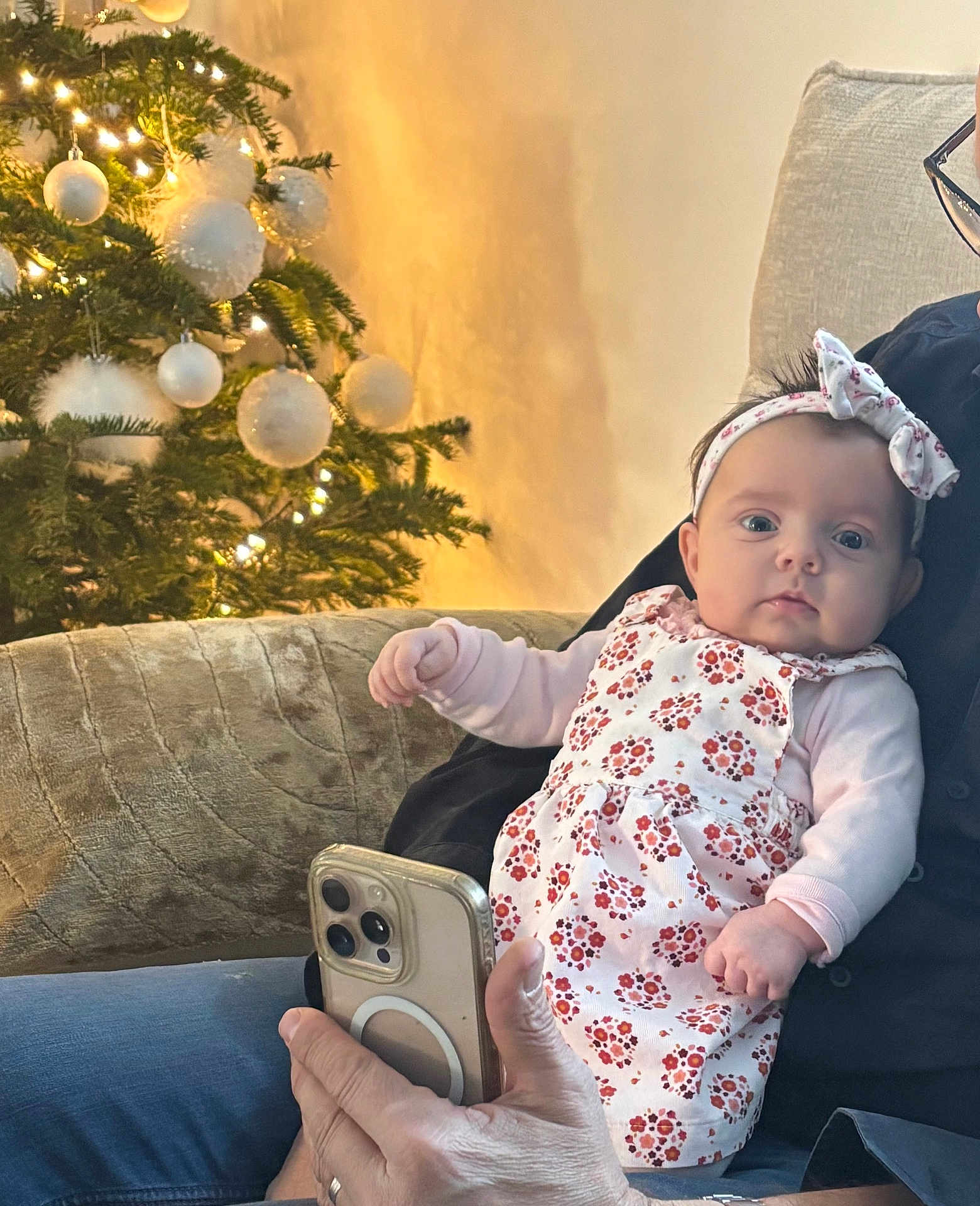Ambre participe au concours pour gagner de l'argent avec cette photo : baby, infant, child, person, adult_hand, smartphone, couch, christmas_tree, ornaments, holiday_lights, headband, dress, floral_pattern, indoor, festive, cozy, seated, warm_lighting, family, portrait
