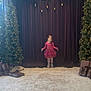 child, girl, pink_dress, curtain, christmas_tree, holiday_decor, presents, carpet, indoor, smile, pose, festive, lights, gift_wrap, stage, young, happy, celebration, event, decorations