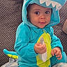 baby, blanket, blue_clothing, child, costume, couch, cozy, cute, dinosaur_onesie, eyes, face, hand, home, hood, indoor, person, plush, seated, smiling, toddler