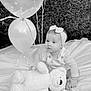 baby, infant, child, balloons, teddy_bear, plush_toy, headband, blanket, outdoor, hedge, grass, portrait, sitting, cute, gaze, ribbon, party, celebration, black_and_white, photography