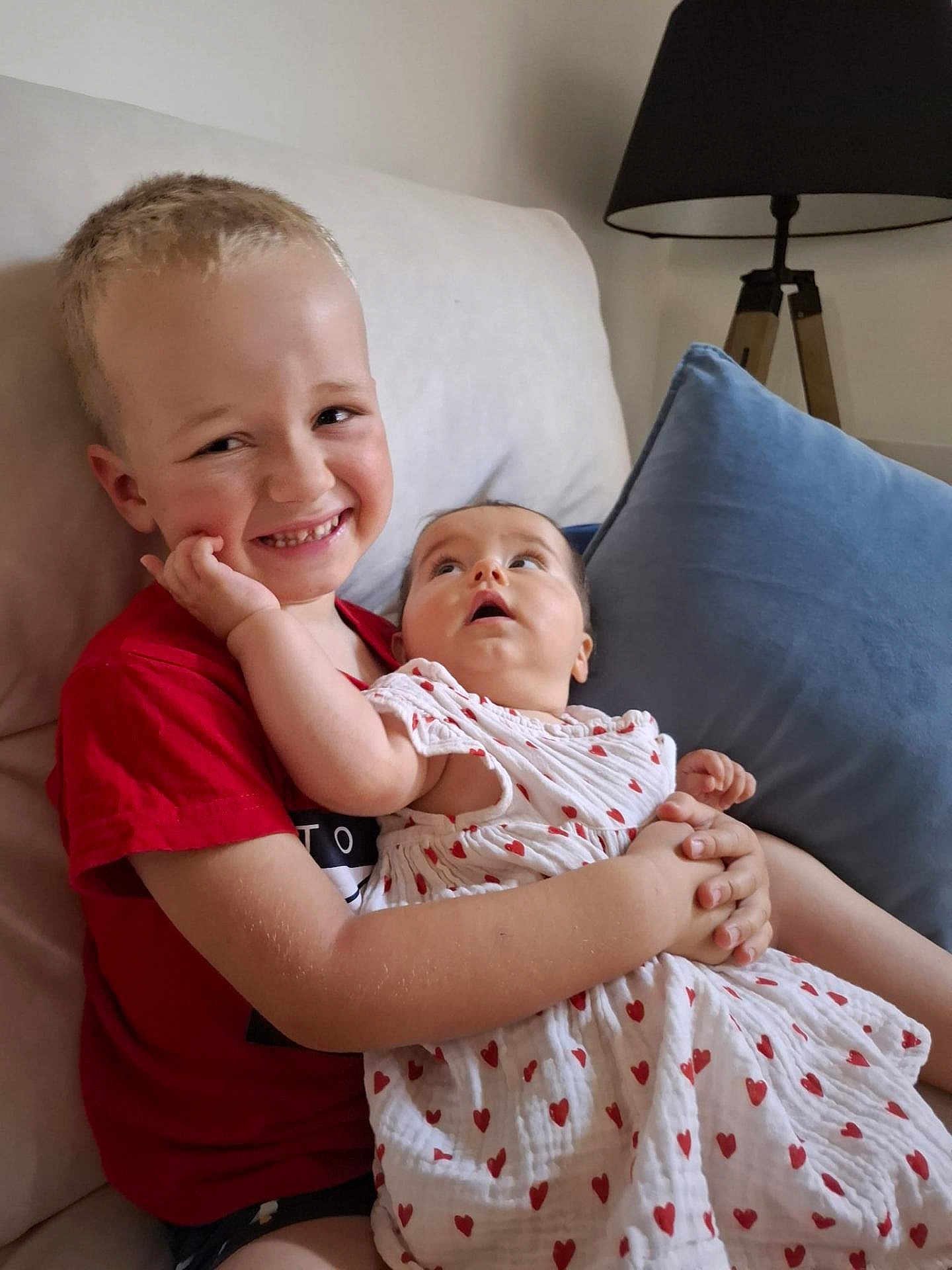 Simon a rejoint le concours — aidez-le/la à gagner de superbes lots ! child, baby, smiling, couch, pillow, lamp, red_shirt, white_dress, hearts_pattern, indoor, brother, sibling, happy, holding, cute, family, portrait, home, seated, love