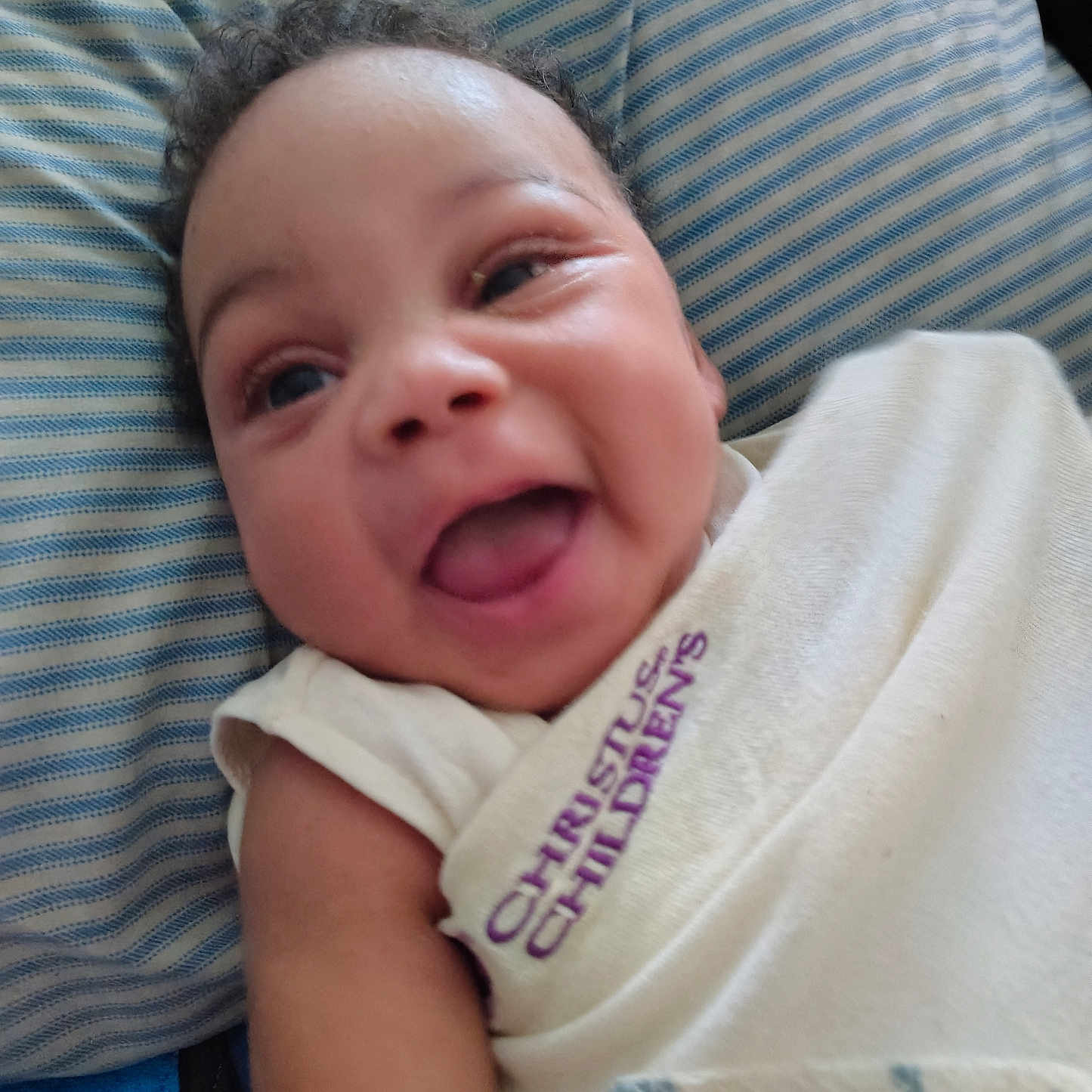 Brandyn Joseph Jr joined the competition — help win amazing prizes! baby, smiling, happy, face, infant, curly_hair, pillow, striped, clothing, white_cloth, text, closeup, portrait, skin, person, cute, tongue_out, lying_down, child, indoors