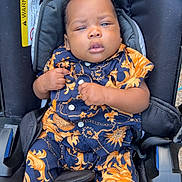 Seven is registered to the contest to win money with this photo: baby, child, car_seat, clothing, patterned_outfit, blue_socks, infant, seatbelt, black_car_seat, cute, portrait, person, indoor, expression, hands, footwear, young_child, sitting, face, young_baby