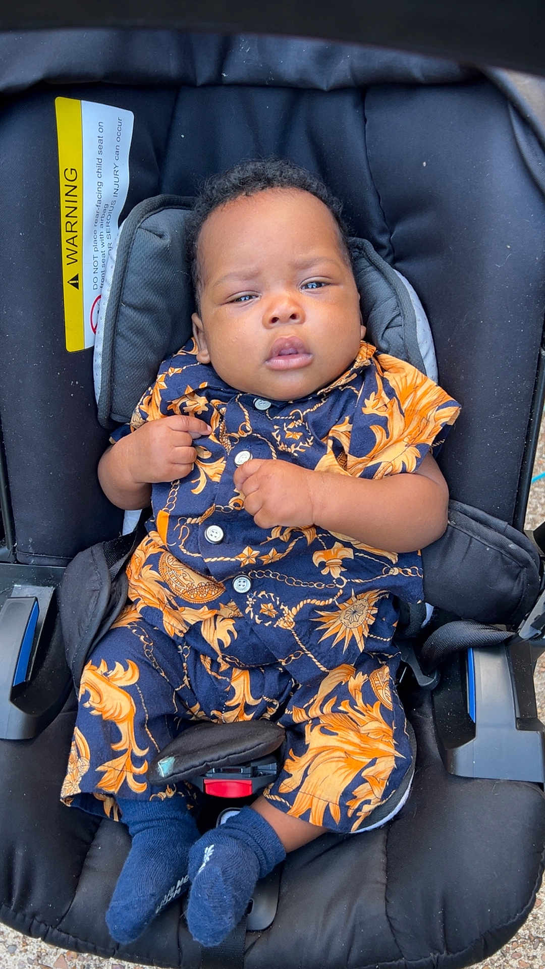 Seven is registered to the contest to win money with this photo: baby, child, car_seat, clothing, patterned_outfit, blue_socks, infant, seatbelt, black_car_seat, cute, portrait, person, indoor, expression, hands, footwear, young_child, sitting, face, young_baby
