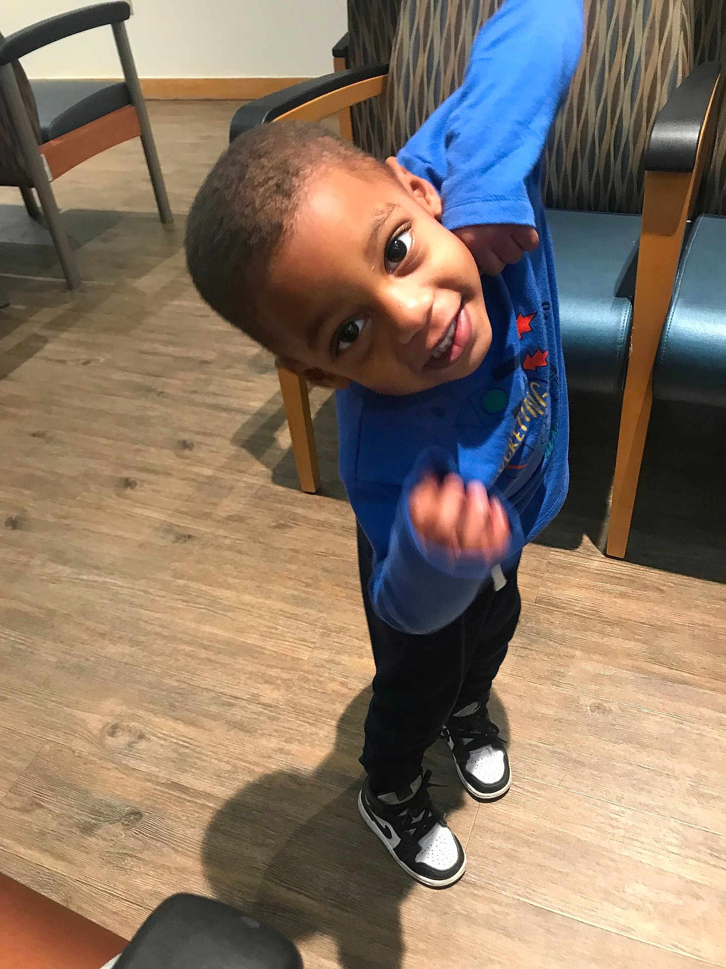 Aiden is registered to the contest to win money with this photo: child, boy, blue_shirt, black_pants, sneakers, indoor, wooden_floor, chair, shadow, smile, playful, pose, happy, young, person, flooring, casual, furniture, head, eyes