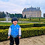 black_pants, blue_jacket, boy, casual_wear, chateau, child, cloudy_sky, daytime, fountain, garden, grass, hedges, historic_building, outdoor, pathway, portrait, smiling, sneakers, standing, young