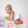 baby, child, crawling, white_dress, headband, pink_flower, large_flowers, pastel_colors, curious_expression, studio_photo, indoor, portrait, cute, infant, flower_decoration, soft_lighting, white_background, kneeling, young_child, adorable