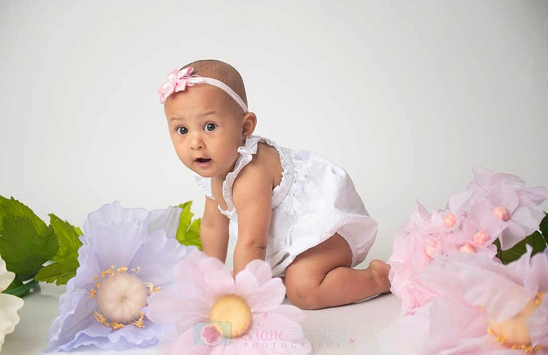 Maria Luiza is registered to the contest to win money with this photo: baby, child, crawling, white_dress, headband, pink_flower, large_flowers, pastel_colors, curious_expression, studio_photo, indoor, portrait, cute, infant, flower_decoration, soft_lighting, white_background, kneeling, young_child, adorable