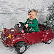 Martin a rejoint le concours — aidez-le/la à gagner de superbes lots ! baby, child, toy_car, green_sweater, gray_cap, smiling, indoor, christmas_tree, winter, holiday, festive, playful, portrait, seat, wheel, vehicle, happy, cute, background, decor