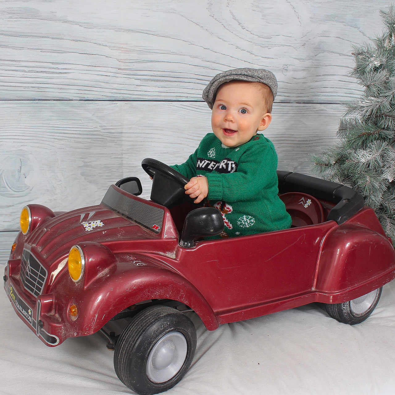 Martin a rejoint le concours — aidez-le/la à gagner de superbes lots ! baby, background, child, christmas_tree, cute, decor, festive, gray_cap, green_sweater, happy, holiday, indoor, playful, portrait, seat, smiling, toy_car, vehicle, wheel, winter