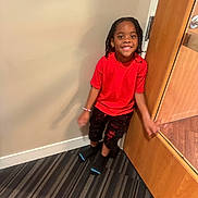 Jahzi is registered to the contest to win money with this photo: child, boy, smile, red_shirt, black_pants, standing, door, indoor, carpet, striped_carpet, wall, happy, person, hair, young, casual_clothing, feet, socks, playful, portrait