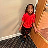 child, boy, smile, red_shirt, black_pants, standing, door, indoor, carpet, striped_carpet, wall, happy, person, hair, young, casual_clothing, feet, socks, playful, portrait