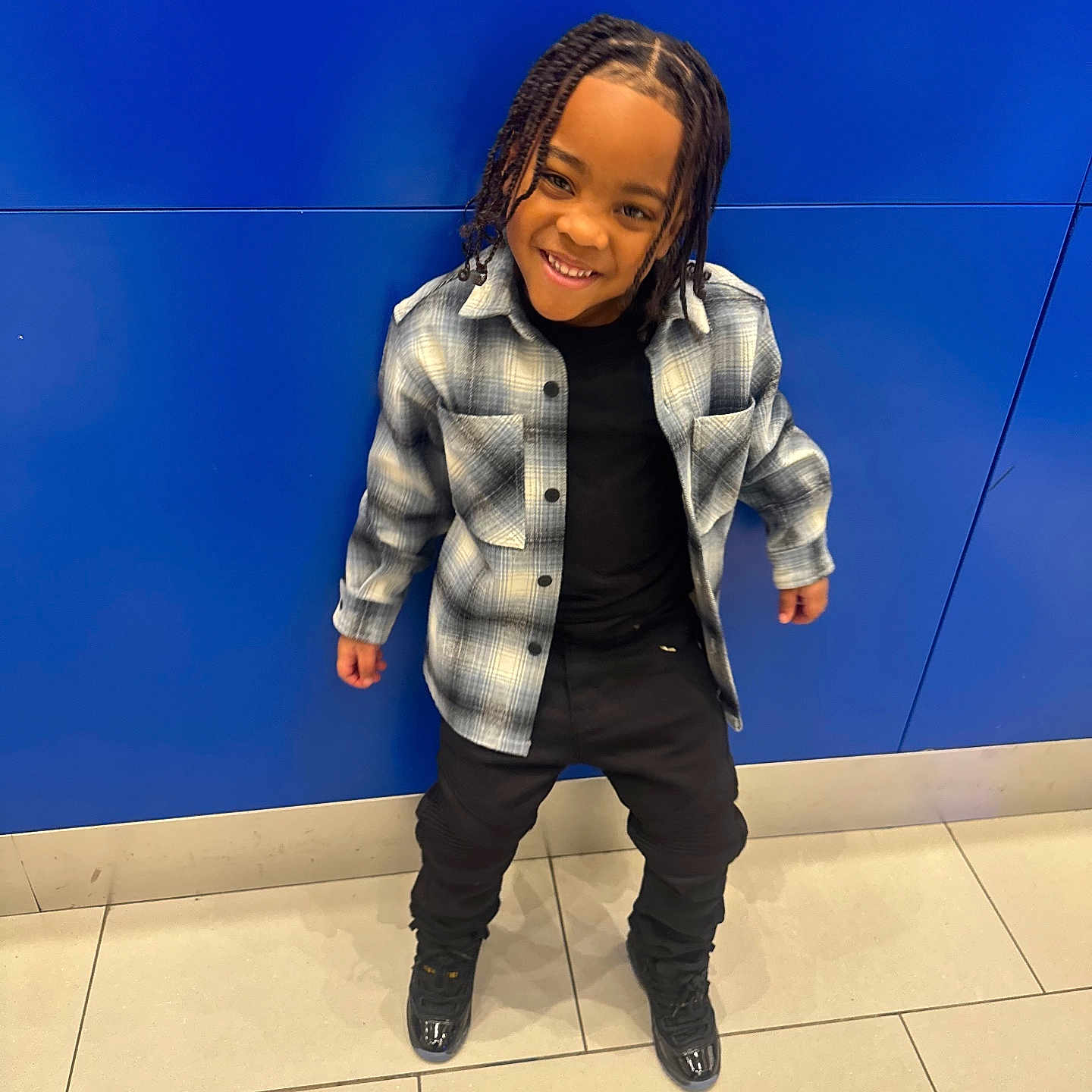 Jahzi is registered to the contest to win money with this photo: child, smiling, braided_hair, plaid_shirt, black_clothing, standing, blue_wall, tile_floor, happy, casual_wear, indoors, person, fashion, portrait, cute, youth, footwear, urban, style, posing