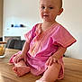 baby, child, pink_clothing, sitting, wooden_table, indoor, portrait, cute, curious, blue_eyes, golden_embroidery, barefoot, infant, hands, legs, face, soft_light, casual, homely, smile