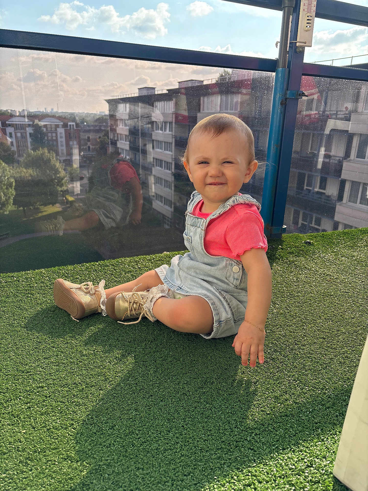 Lyna participe au concours pour gagner de l'argent avec cette photo : toddler, child, sitting, overalls, pink_shirt, shoes, outdoor, balcony, glass_railing, reflection, urban, buildings, sky, clouds, grass, daylight, portrait, cute, playful, sunlight
