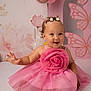 baby, toddler, pink_dress, flower_decoration, barefoot, smiling, happy, sitting, curly_hair, hair_accessories, balloons, butterfly_wall_art, pastel_colors, indoor, child, portrait, cute, celebration, decor, playful