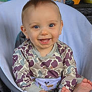 David is registered to the contest to win money with this photo: baby, child, smiling, blue_eyes, sitting, clothing, patterned_outfit, foot, chair, indoor, happy, face, young_child, cute, person, home, decor, colorful, toy, expression