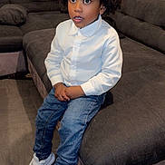 Edgar participe au concours pour gagner de l'argent avec cette photo : child, afro_hair, white_shirt, blue_jeans, white_sneakers, sofa, living_room, curious_expression, indoor, seated, floor_tiles, casual_clothing, young_boy, portrait, furniture, comfortable, modern, home, cute, fashion