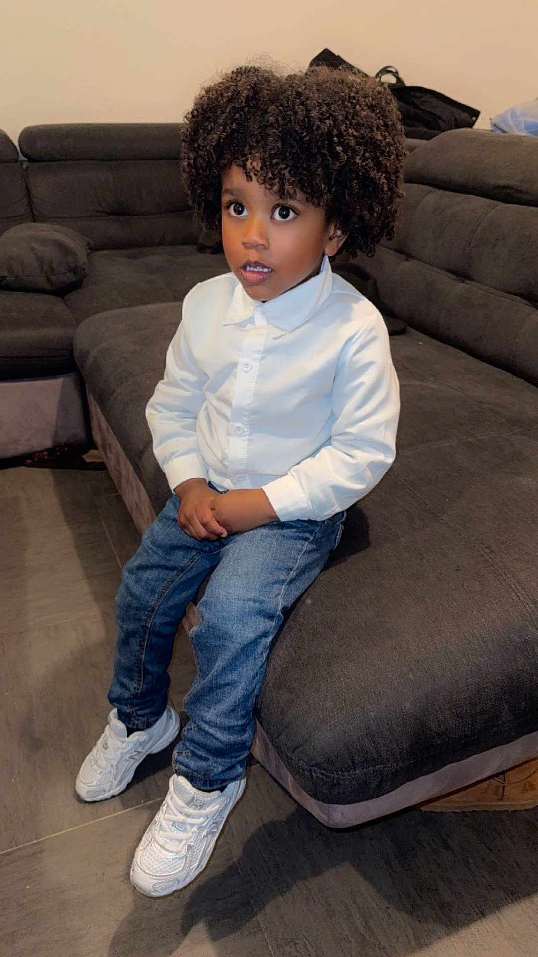 Edgar participe au concours pour gagner de l'argent avec cette photo : child, afro_hair, white_shirt, blue_jeans, white_sneakers, sofa, living_room, curious_expression, indoor, seated, floor_tiles, casual_clothing, young_boy, portrait, furniture, comfortable, modern, home, cute, fashion