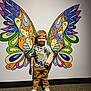 child, boy, butterfly_wings, mural, wall_art, cast, arm_cast, standing, indoor, carpet, tshirt, pants, sneakers, smiling, colorful, playful, young_child, portrait, casual_clothing, artistic_background