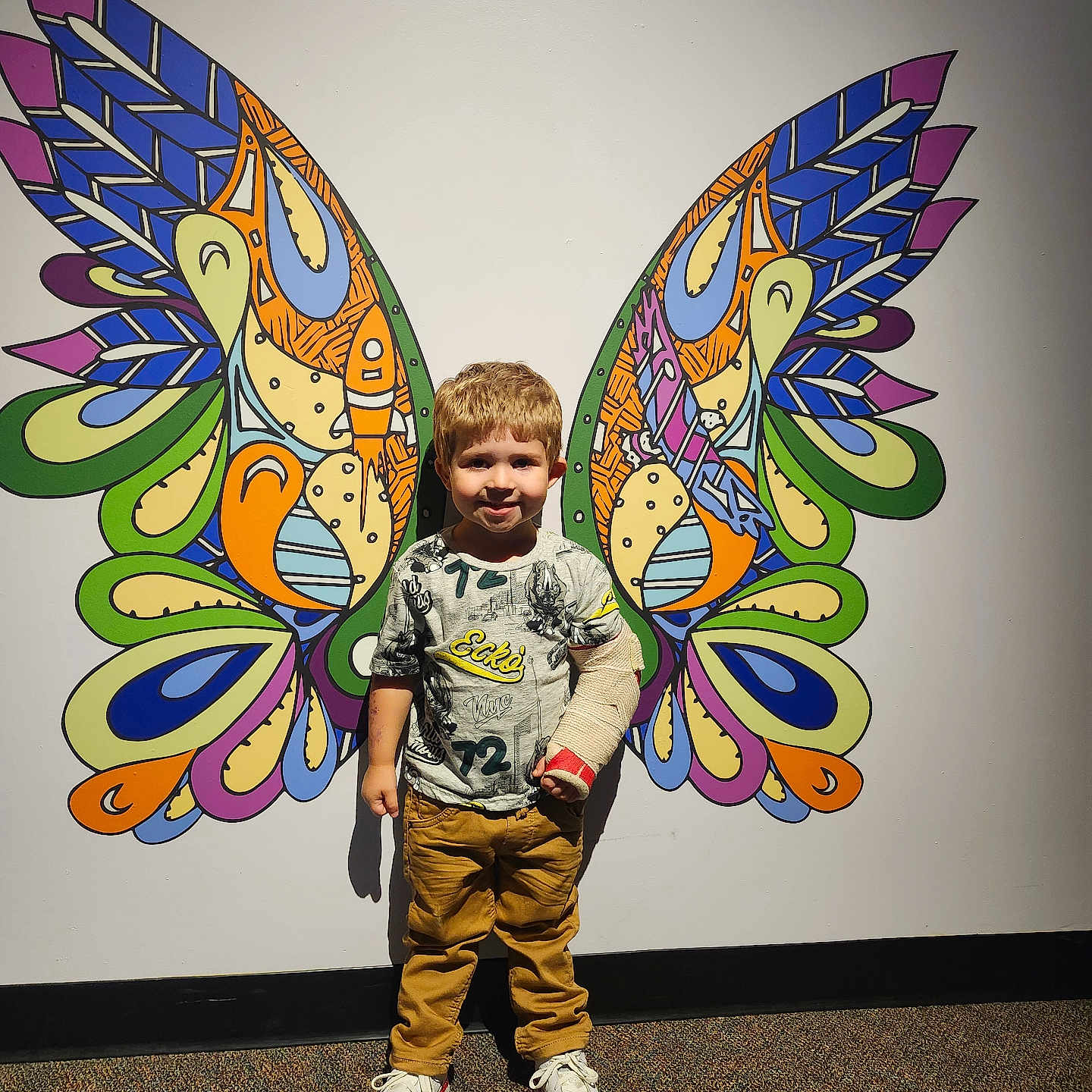 Bentley is registered to the contest to win money with this photo: arm_cast, artistic_background, boy, butterfly_wings, carpet, cast, casual_clothing, child, colorful, indoor, mural, pants, playful, portrait, smiling, sneakers, standing, tshirt, wall_art, young_child