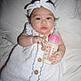 baby, infant, bottle, milk, white_dress, headband, bed, blanket, sleeping_area, wooden_buttons, hand, face, eyes, curious, lying_down, cute, indoors, skin, person, child