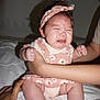 baby, crying, infant, hands, clenched_fists, headband, floral_pattern, pink_clothing, white_bed, adult_hands, skin, expression, indoor, shadow, person, cute, chubby, legs, toes, closeup