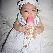 Aya is registered to the contest to win money with this photo: baby, child, bottle, feeding, headband, white_clothing, embroidery, bed, blanket, infant, cute, portrait, lying_down, hand, feet, person, indoors, soft_lighting, smile, milk