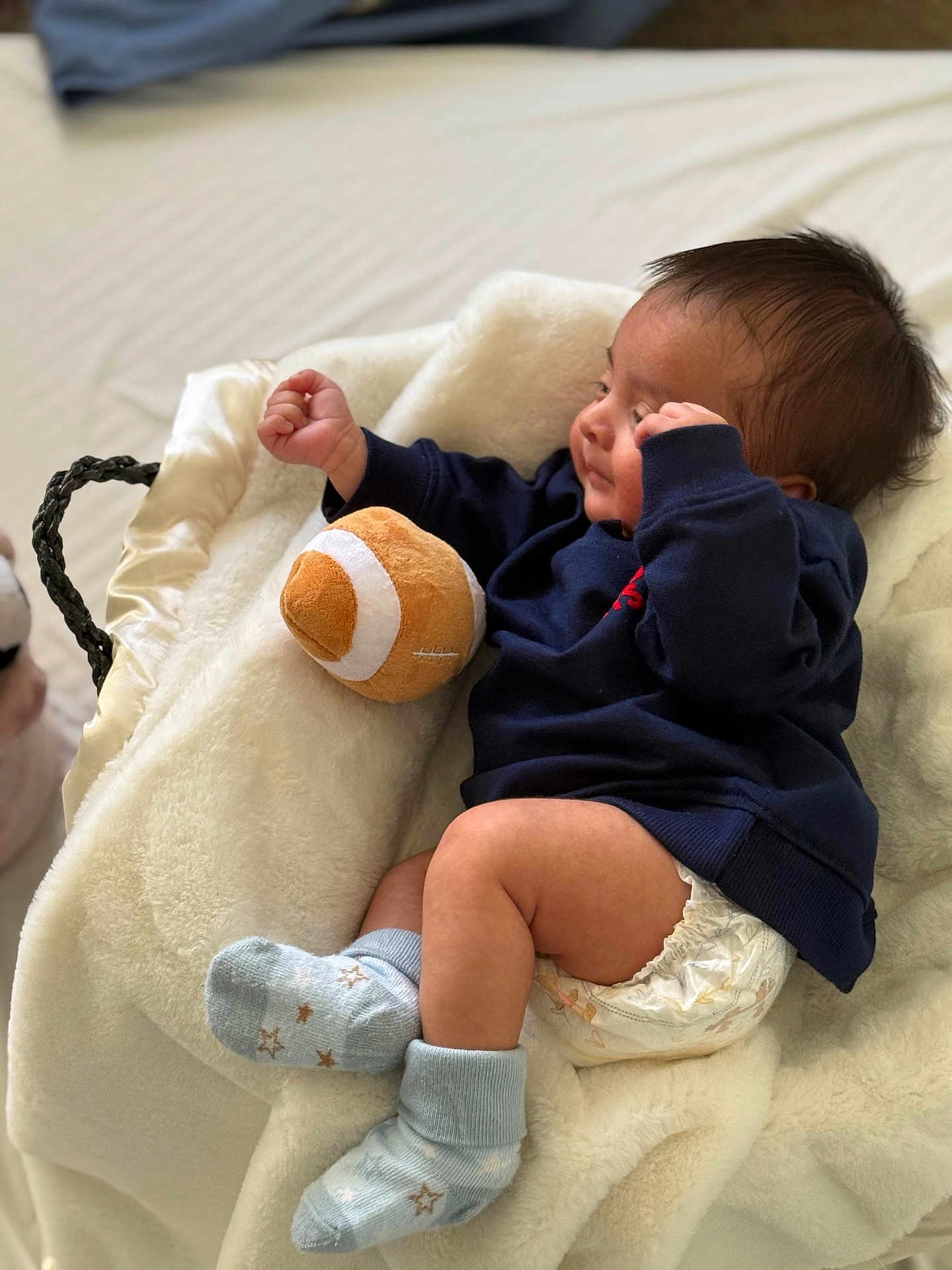 Idris is registered to the contest to win money with this photo: baby, infant, newborn, diaper, socks, plush_football, basket, blanket, cozy, lying_down, side_profile, hair, hand, arm, clothing, bed, soft_toy, portrait, adorable, nursery