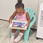 Gracy a rejoint le concours — aidez-le/la à gagner de superbes lots ! child, girl, pink_clothing, sitting, plastic_chair, photo_frame, floor, indoor, curly_hair, socks, toy, light_wall, wood_floor, looking_down, holding, curiosity, young_child, casual, home, innocence