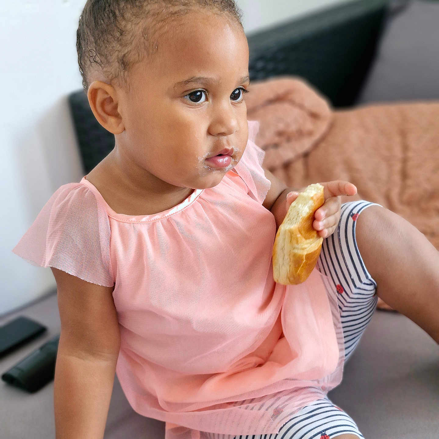 Gracy a rejoint le concours — aidez-le/la à gagner de superbes lots ! bread, casual, child, couch, crumbs, curious, expression, face, food, girl, hair_bun, hand, indoor, pink_dress, portrait, relaxed, sitting, skin, striped_leggings, toddler