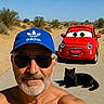 man, shirtless, blue_cap, sunglasses, black_cat, animated_car, desert, road, sunny, outdoor, selfie, smile, beard, gray_hair, vehicle, nature, clear_sky, relaxing, funny, cartoon