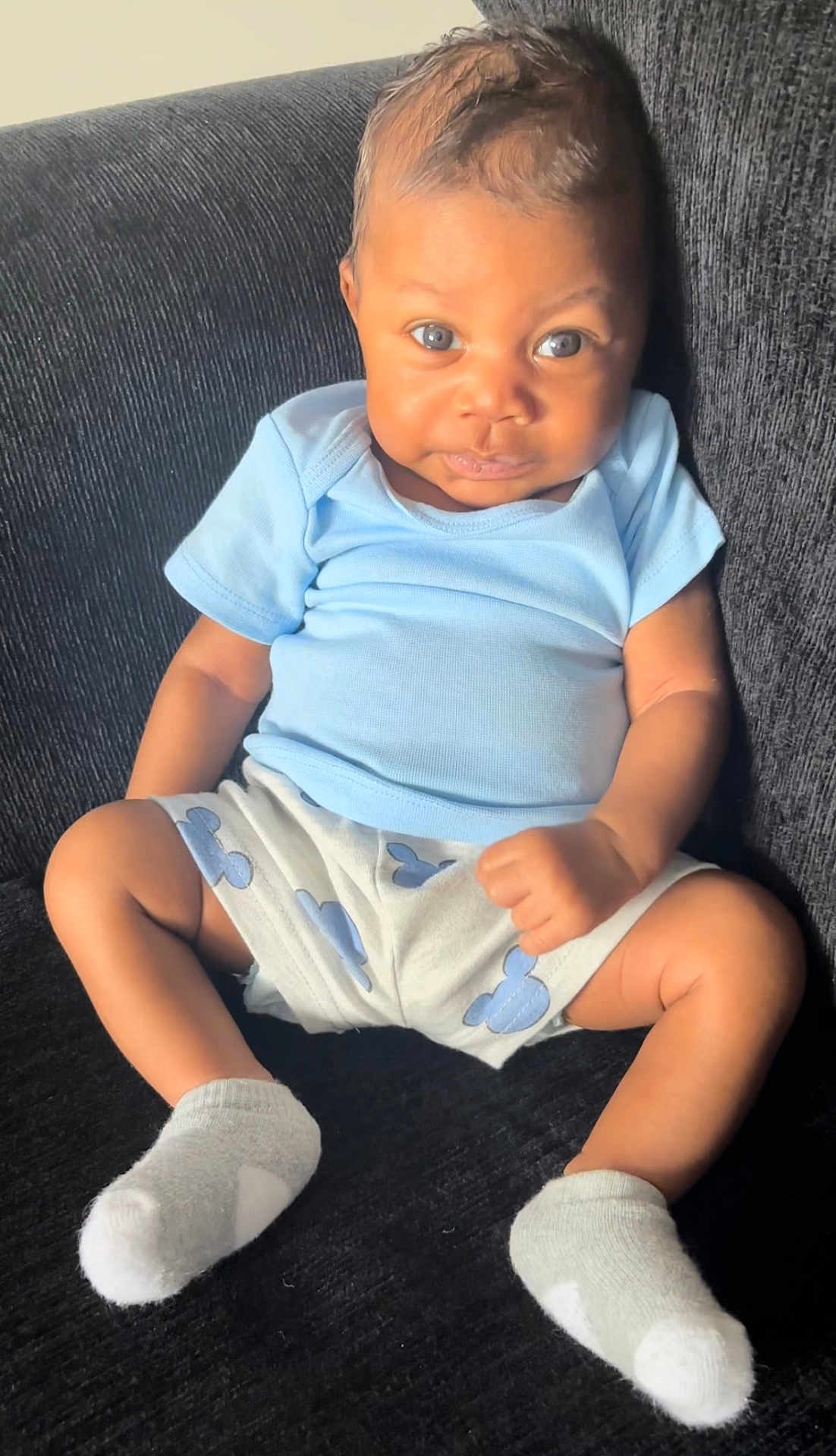 Zaiden is registered to the contest to win money with this photo: baby, infant, child, blue_shirt, shorts, socks, couch, furniture, seated, portrait, face, eyes, skin, expression, indoor, cute, person, young_child, sitting, clothing