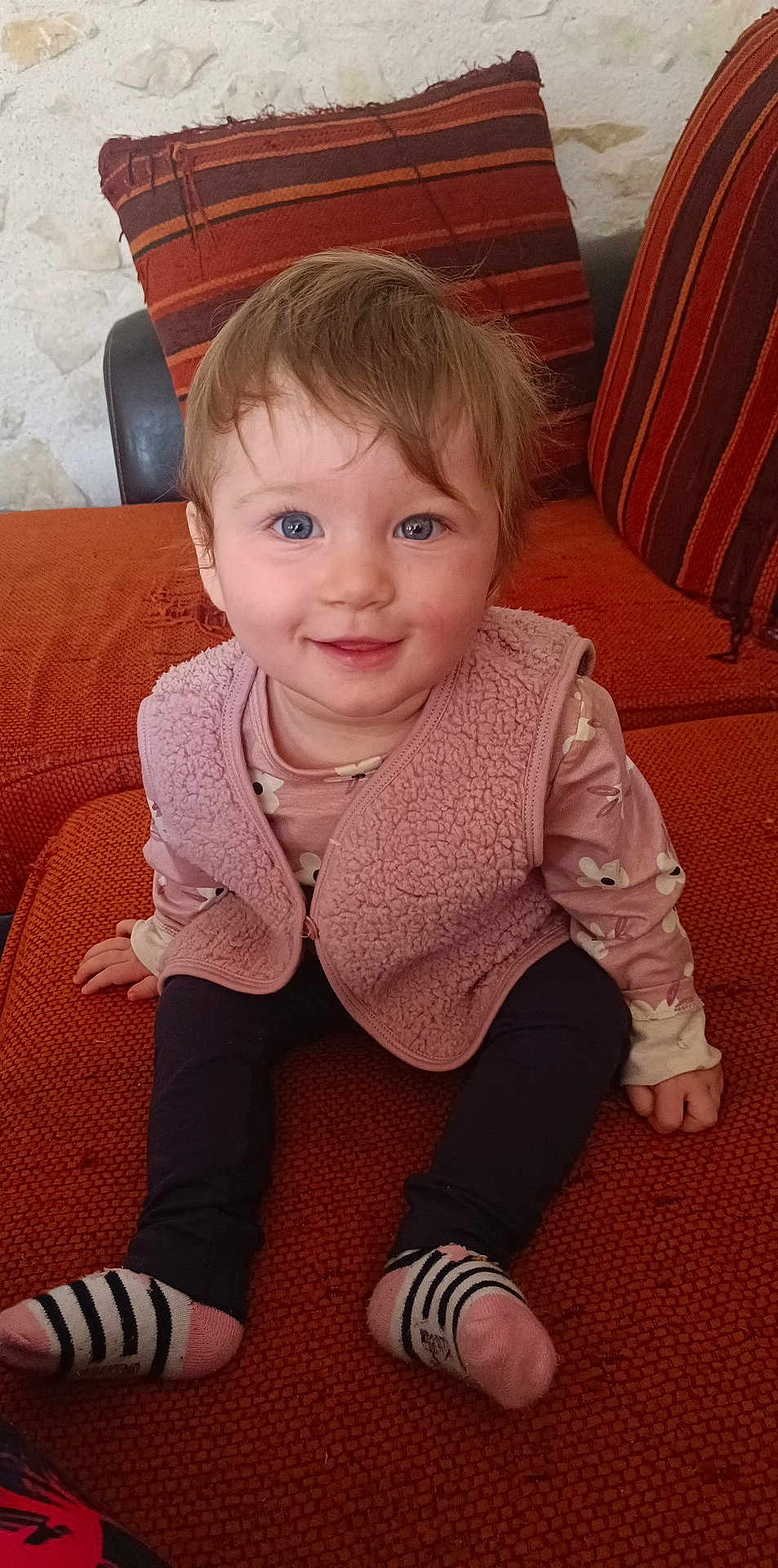 Léonie participe au concours pour gagner de l'argent avec cette photo : toddler, child, smiling, blue_eyes, pink_vest, floral_shirt, striped_socks, orange_couch, indoor, happy, sitting, cute, young_child, hair, face, person, cushion, furniture, cozy, home