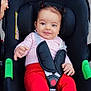 baby, infant, stroller, child, smiling, red_pants, white_socks, hair_pigtails, car_seat, safety_harness, outdoor, sidewalk, cute, happy, person, small_hands, bright_clothing, seatbelt, playful, toddler