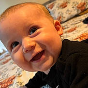 Myles joined the competition — help win amazing prizes! baby, bedspread, black_clothing, cheeks, child, closeup, cute, eyes, face, floral_pattern, happy, head, indoors, infant, lying_down, person, portrait, skin, smiling, young_child