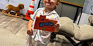 Hadley is registered to the contest to win money with this photo: toddler, child, pajamas, barefoot, carpet, candy_box, runts, rocking_horse, traffic_cone, television, living_room, furniture, wood_floor, cute, expression, toy, indoor, person, smiling, home