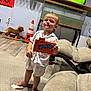 toddler, child, pajamas, barefoot, carpet, candy_box, runts, rocking_horse, traffic_cone, television, living_room, furniture, wood_floor, cute, expression, toy, indoor, person, smiling, home