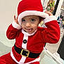 toddler, child, santa_claus_costume, red_clothing, hat, playful, tongue_out, indoor, portrait, cute, holiday, festive, smiling, person, face, cartoon_poster, background, furniture, belt, holiday_season
