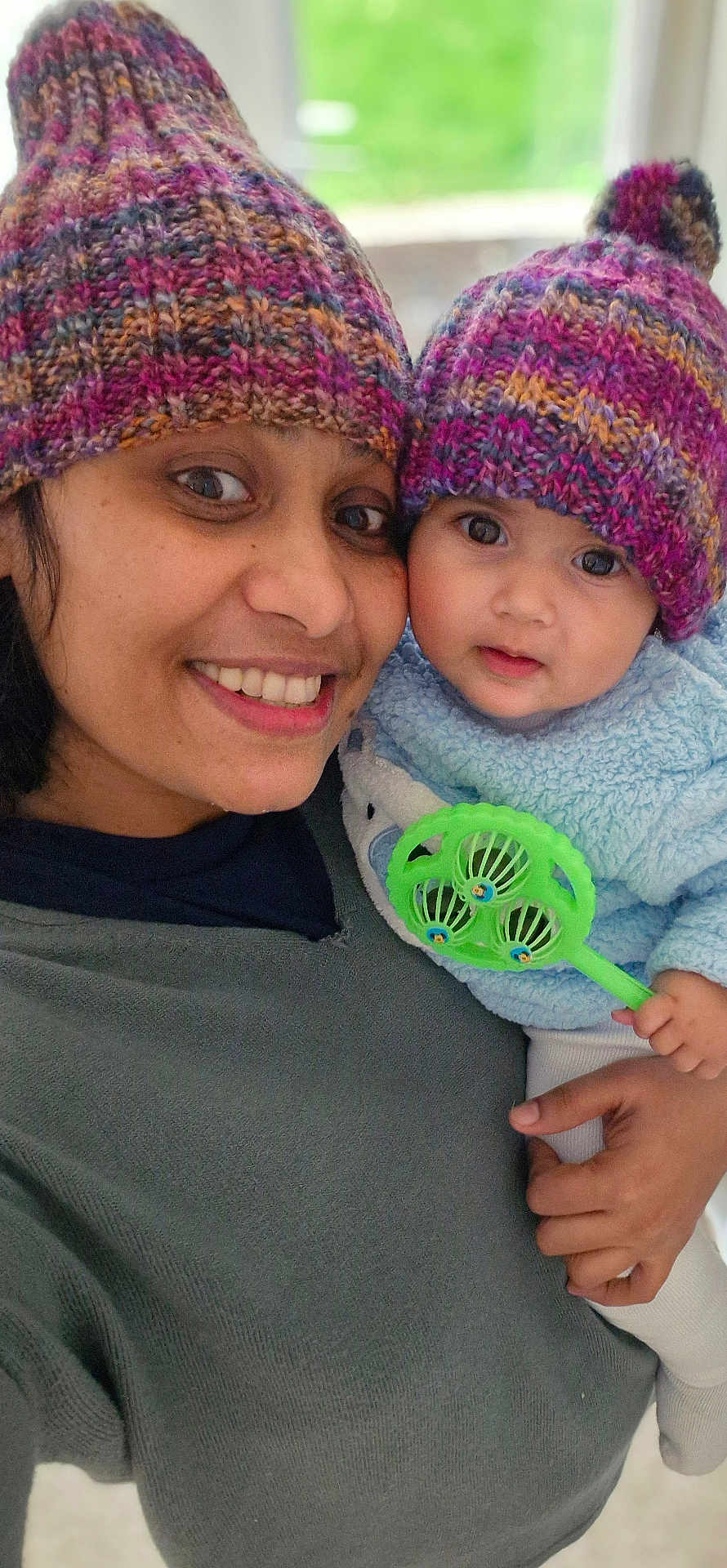 Nishitha is registered to the contest to win money with this photo: adult, baby, child, close_up, colorful_hat, cute, face, family, green_toy, hand, happy, holding, indoor, knitted_hat, person, portrait, smiling, sweater, toy, warm_clothing