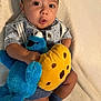 baby, child, plush_toy, cookie_monster, stuffed_animal, yellow_cookie, blue_socks, patterned_shirt, cute, wide_eyes, surprised_expression, baby_clothes, soft_blanket, indoors, portrait, cuddly, comfort, sitting, toy, infant