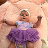 baby, infant, child, pink_cushion, purple_tutu, headband, onesie, toy, indoor, soft_texture, cute, lying_down, arms_raised, colorful_clothing, white_socks, carpet, happy_expression, soft_lighting, playful, portrait
