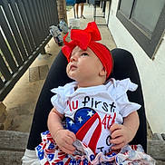 Avery is registered to the contest to win money with this photo: baby, infant, red_bow, patriotic_clothing, porch, person, man, outdoor, daylight, headband, skirt, socks, looking_up, seated, hand, white_shirt, black_pants, wooden_bench, house, railing
