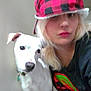 Lucki joined the competition — help win amazing prizes! person, dog, white_dog, hat, red_and_black_hat, blonde_hair, black_shirt, indoor, pet, collar, portrait, close_up, casual, looking_at_camera, companion, animal, friendship, candid, soft_lighting, plain_background