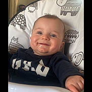 Phillip is registered to the contest to win money with this photo: baby, child, high_chair, smile, happy, indoor, animal_print, toddler, face, blue_eyes, clothing, feeding_chair, cute, infant, hand, cheeks, light, window, room, portrait