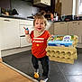 Elio participe au concours pour gagner de l'argent avec cette photo : toddler, child, kitchen, egg_carton, orange_shirt, smiling, standing, floor, toy, ball, cabinet, countertop, appliance, window, indoor, person, clothing, pants, socks, happy