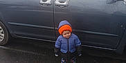 Lucas is registered to the contest to win money with this photo: child, winter_clothing, jacket, beanie, boots, car, snow, parking_lot, wet_asphalt, outdoor, cold_weather, vehicle, pavement, road_marking, hood, young_child, standing, casual_wear, daylight, person
