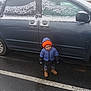 child, winter_clothing, jacket, beanie, boots, car, snow, parking_lot, wet_asphalt, outdoor, cold_weather, vehicle, pavement, road_marking, hood, young_child, standing, casual_wear, daylight, person