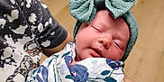 Serenity is registered to the contest to win money with this photo: newborn, baby, sleeping, headband, bow, blanket, floral, swaddle, person, hand, indoor, wooden_floor, door, patterned_clothing, closeup, infant, peaceful, human, holding, portrait