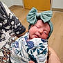 newborn, baby, sleeping, headband, bow, blanket, floral, swaddle, person, hand, indoor, wooden_floor, door, patterned_clothing, closeup, infant, peaceful, human, holding, portrait